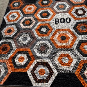 May include: A quilt featuring a repeating hexagonal pattern in orange, black, white, and gray fabrics. The design includes various patterned textiles with motifs like spiders and webs. The word BOO is printed in black letters on a white hexagon.