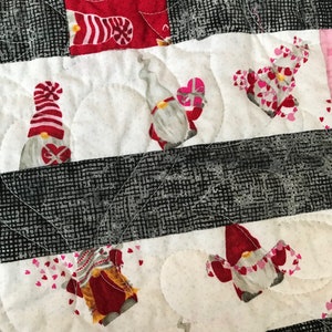 May include: A white and red patchwork quilt with a pattern of gnomes and hearts. The gnomes are wearing red hats and have white beards. The hearts are pink and white.