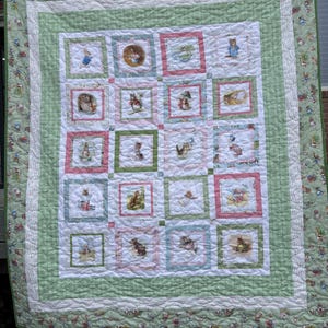 May include: A handmade quilt featuring a patchwork design with squares showcasing illustrations of rabbits and other animals. The quilt has a white center with pink and green borders, and a green patterned edge. The quilt is hanging outside.