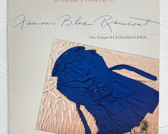 Könnte beinhalten: Ein Schallplattencover mit dem Text Jennifer Warnes Famous Blue Raincoat The Songs of Leonard Cohen. Das Bild zeigt einen leuchtend blauen Trenchcoat, der flach auf einem hellen pfirsichfarbenen Stoffuntergrund liegt, neben einem kleinen blauen Objekt.