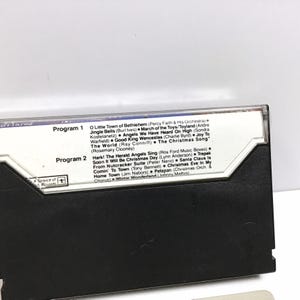 May include: A black cassette tape with a white label. The label has the text "Program 1" and "Program 2" with a list of Christmas songs under each program. The cassette tape is a vintage item.
