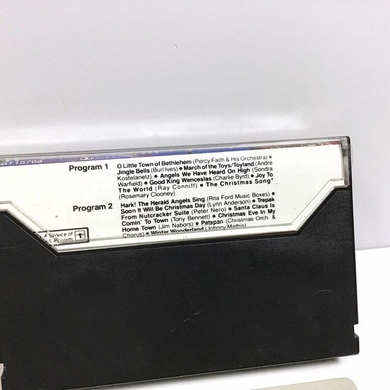May include: A black cassette tape with a white label. The label has the text "Program 1" and "Program 2" with a list of Christmas songs under each program. The cassette tape is a vintage item.