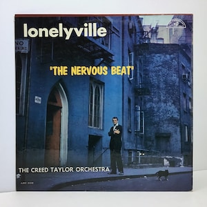 May include: A vintage vinyl record album cover for "Lonelyville" featuring "The Nervous Beat" by The Creed Taylor Orchestra. The cover art depicts a man in a suit standing on a city street with a blue building backdrop and a black cat.