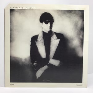 May include: Black and white album cover featuring T Bone Burnett. Burnett is wearing sunglasses and a black suit with a decorative lapel. The album cover has the text "T Bone Burnett" at the top and "M.M.M." at the bottom.
