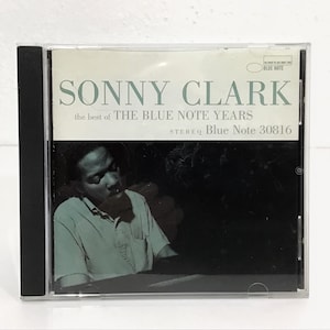 May include: A CD case featuring the album "Sonny Clark: The Best of The Blue Note Years." The cover has a black and white photo of a man in a light blue shirt. The album title and label information are in white and light blue.