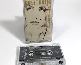 May include: A clear plastic cassette tape case with a black cassette tape inside. The cassette tape has a label with the text "Eurythmics - Savage" and a list of songs. The case has a picture of a woman with blonde hair on the front.