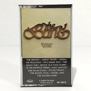 May include: A vintage cassette tape featuring "The Band" logo in brown script. The cassette case is clear, with a beige background and a list of song titles. The Capitol Records logo is visible.