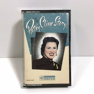 May include: A black cassette tape case with a photo of Patsy Cline on the front. The title of the cassette is "The Patsy Cline Story" and the label is "Original Decca".