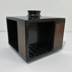 May include: A square-shaped black plastic media storage rack featuring wood-grain side panels and a central circular top opening. The unit has open sides with internal slotted dividers designed for organizing individual items in a grid.
