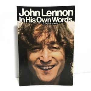 May include: A book cover featuring a close-up photo of John Lennon smiling. The title of the book is "John Lennon In His Own Words. Compiled by Miles."