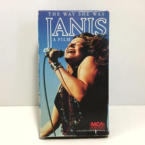 May include: A vintage VHS tape for the film "The Way She Was Janis." The cover features a vibrant image of Janis Joplin singing into a microphone, with the title in bold white letters against a blue sky background. The MCA Home Video logo is visible.
