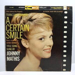 May include: A black and white vinyl record cover featuring a woman with blonde hair and a white shirt. The cover has the title "A Certain Smile" and the text "Music composed by Alfred Newman" and "Title Song Sung By Johnny Mathis".