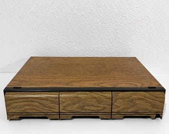 Könnte beinhalten: Ein rechteckiger Schreibtisch-Organizer aus Holz mit einer glatten Oberfläche und drei horizontalen Schubladen mit sichtbarer Holzmaserung. Die Einheit ist mit einem dünnen schwarzen Metallrahmen an der Vorderkante versehen.