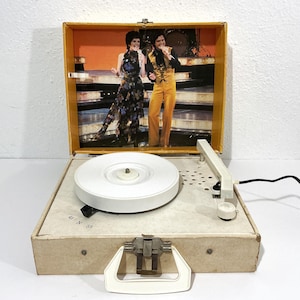 May include: A portable record player with a white turntable and a cream-colored case. The interior lid features a printed image of a singing duo on stage. The control panel includes the text 45 N 33 and a white handle is attached to the front.