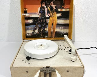 Könnte beinhalten: Ein tragbarer Plattenspieler mit weißem Plattenteller und cremefarbenem Gehäuse. Der Innendeckel zeigt ein aufgedrucktes Bild eines singenden Duos auf der Bühne. Das Bedienfeld enthält den Text 45 N 33 und ein weißer Griff ist vorne angebracht.