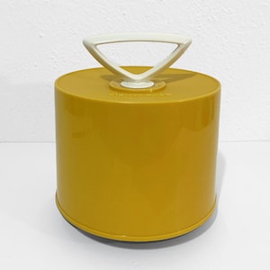 May include: A cylindrical mustard yellow plastic storage container with a smooth finish and a white triangular handle attached to the center of the lid. The container features a solid, opaque body and a distinct, molded geometric design on top.