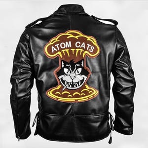 Può includere: Giacca in pelle nera con una grande toppa Atom Cats sul retro. La toppa presenta un gatto dei cartoni animati che fuma una sigaretta, ambientato su un design a fungo atomico con le parole "ATOM CATS".