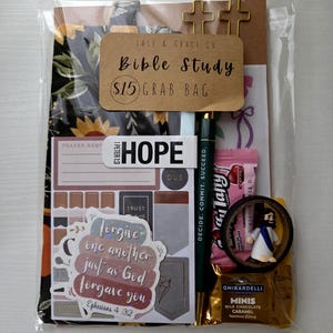 May include: A clear cellophane bag containing a Bible study grab bag. The bag includes a pen, stickers, a chocolate square, a small figurine, and a tag that reads "Bible Study $15 Grab Bag". The bag also contains a floral print item.