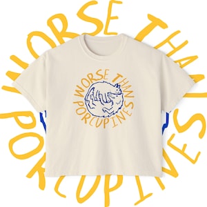 May include: Cream-colored t-shirt with a round neck and short sleeves. The shirt features a blue and orange graphic of a cat curled up inside a circle with the words "Worse Than Porcupines" around it. The words "Worse Than Porcupines" are also written in a circle around the shirt.