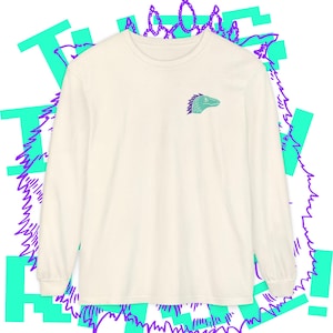 May include: Cream-coloured long-sleeved shirt with a small teal and purple dinosaur head graphic on the chest. The shirt has a crew neck and long sleeves. The background features teal and purple abstract shapes.