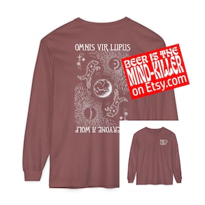 May include: A long-sleeve shirt in a muted red-brown color. The back features a white graphic with celestial elements and the text "OMNIS VIR LUPUS" and "EVERYONE A WOLF". A red sticker with the text "BEER IS THE MIND-KILLER on Etsy.com" is visible.