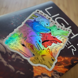 Peut inclure: Autocollant holographique repr&eacute;sentant une meute de t&ecirc;tes de loup stylis&eacute;es de diff&eacute;rentes couleurs, dont le rose, le bleu et le vert. L'autocollant est plac&eacute; sur un livre avec le mot "LIGHT" visible. L'autocollant a une finition brillante et iris&eacute;e.