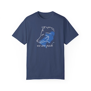 以下が含まれることがあります： 白い輪郭の狼と人間の顔が重なり合ったネイビーブルーのTシャツ。画像の下に「we are pack」というテキストが印刷されています。