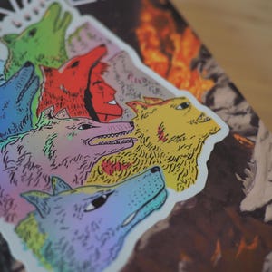 Peut inclure: Un autocollant repr&eacute;sentant une meute de t&ecirc;tes de loups stylis&eacute;es de diff&eacute;rentes couleurs, dont le vert, le rouge, le jaune et le bleu, avec un &eacute;clat holographique. L'autocollant est plac&eacute; sur un livre avec le mot "ANGER" visible.