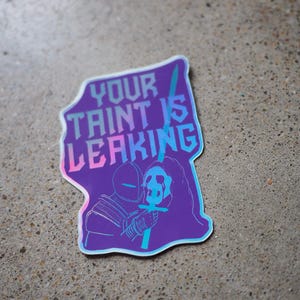 以下が含まれることがあります： 「YOUR TAINT IS LEAKING」の文字が入った紫色のステッカー。十字型のオブジェクトを持つ騎士のような人物が描かれています。デザインはホログラフィック効果があります。