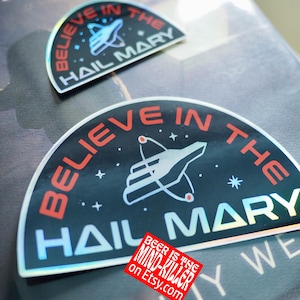 Puede incluir: Dos pegatinas semicirculares con el texto "BELIEVE IN THE HAIL MARY" en rojo y blanco. Las pegatinas presentan un diseño de nave espacial y estrellas sobre un fondo oscuro. Una pequeña pegatina cuadrada roja dice "BEER IS THE MIND-KILLER on Etsy.com".