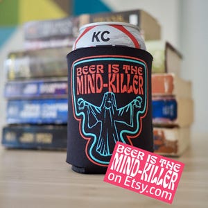 Puede incluir: Enfriador de latas de cerveza negro con el texto "BEER IS THE MIND-KILLER" en rojo y turquesa. El enfriador tiene un gráfico de una figura fantasmal. Una pegatina rosa con el mismo texto y Etsy.com está en primer plano.