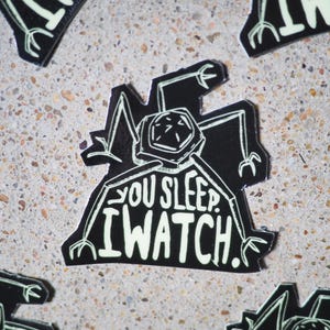 May include: Black and white sticker with a robot-like creature with the text "You sleep. I watch."