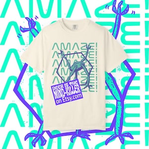 Puede incluir: Camiseta color crema con un diseño gráfico que presenta una figura alienígena estilizada dentro de un hexágono, con la palabra "AMAZE!" repetida verticalmente. Un rectángulo morado en la parte inferior dice "BEER IS THE MIND-KILLER on Etsy.com".