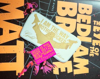 May include: A book cover with the title "The Eye of the Bedlam" in orange letters on a black background. A sticker with the text "I AM THE WAY MOTHERFUCKER" and a pink sticker that says "BEER IS THE MIND-KILLER on etsy.com" are on the cover.
