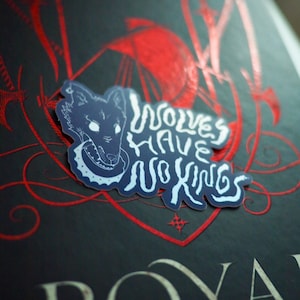 Puede incluir: Una pegatina con la cabeza de un lobo azul y las palabras blancas "Wolves Have No Kings". La pegatina está sobre un libro con detalles decorativos rojos y la palabra "ROYAL" en blanco.