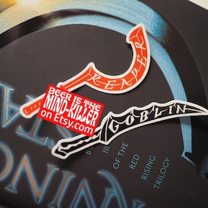 May include: Two stickers on a book. One is a red and white curved design with the word "REAPER". The other is a black and white design with the word "GOBLIN". A red rectangle sticker reads "BEER IS THE MIND-KILLER on Etsy.com".