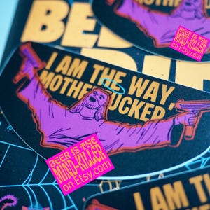 May include: A collection of stickers with a black background and yellow and purple text. The stickers feature a cartoon image of Jesus holding a gun, with the text "I AM THE WAY MOTHERFUCKED". A pink sticker reads "BEER IS THE MIND-KILLER on Etsy.com".