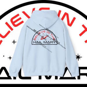 以下が含まれることがあります： 背中にグラフィックが入った水色のパーカー。「Believe in the Hail Mary」と書かれており、赤と白のデザインと黒い文字で「Hail Mary」と書かれています。パーカーにはフードと長袖が付いています。