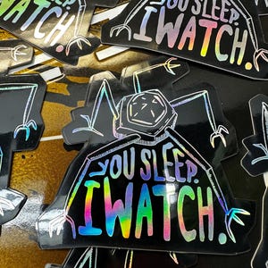 May include: A black and white sticker with a holographic finish. The sticker features a cartoon creature with the text "You Sleep I Watch!" in a rainbow holographic font.