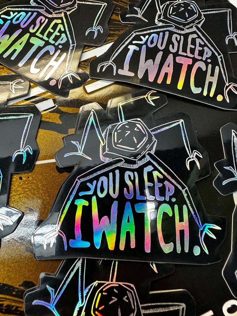May include: A black and white sticker with a holographic finish. The sticker features a cartoon creature with the text "You Sleep I Watch!" in a rainbow holographic font.