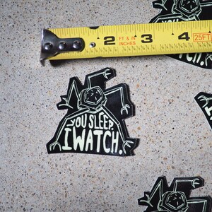 May include: Black and white sticker with a robot-like creature and the text "You sleep. I watch."