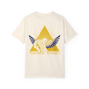 May include: A cream colored t-shirt with a gold and blue graphic of a winged creature inside a triangle. The text "Morning Knight" is printed below the graphic.