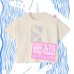 May include: Cream-colored t-shirt with a crew neck and short sleeves. The shirt features a graphic of hands and the word "Beloved" in a script font. A pink rectangle with the text "BEER IS THE MIND-KILLER on Etsy.com" is on the front.