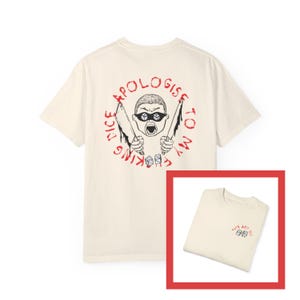 Pode incluir: Uma camiseta bege com uma estampa gráfica nas costas. O gráfico apresenta um personagem de desenho animado com uma faca em cada mão e o texto "Apologise to my dice king". A frente da camiseta tem um pequeno gráfico de dados com o texto "Give and Roll".