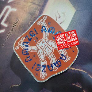 May include: An orange sticker with a white robot-like figure and the words "Amaze!" in blue glitter. A small red rectangle reads "Beer is the Mind-Killer on Etsy.com". The sticker is on a book.