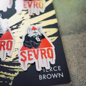 Puede incluir: Una pegatina blanca con un triángulo rojo y una ilustración de lobo negro. El lobo tiene ojos rojos y colmillos. El texto "SEVRO" está escrito en letras rojas que gotean.