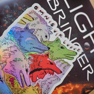 Peut inclure: Un autocollant holographique repr&eacute;sentant une meute de loups color&eacute;s. Les loups sont repr&eacute;sent&eacute;s dans des tons de bleu, vert, rouge et violet. Le texte "LIGHT BRINGER" et "OMNIS VIR LUPUS" est pr&eacute;sent.