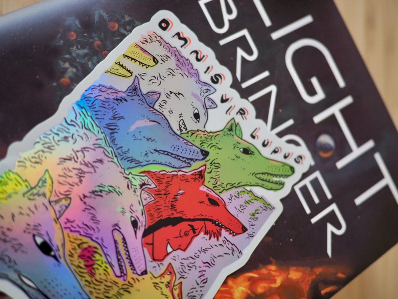 Peut inclure: Un autocollant holographique repr&eacute;sentant une meute de loups color&eacute;s. Les loups sont repr&eacute;sent&eacute;s dans des tons de bleu, vert, rouge et violet. Le texte "LIGHT BRINGER" et "OMNIS VIR LUPUS" est pr&eacute;sent.