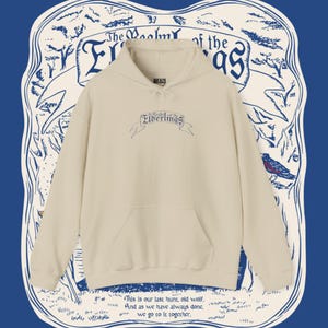 Op de afbeelding: Crèmekleurige hoodie met een voorzak en een capuchon met trekkoord. De tekst "The Balm of the Elderlings" is op de borst geborduurd. De hoodie staat tegen een blauwe achtergrond met decoratieve tekst en illustraties.