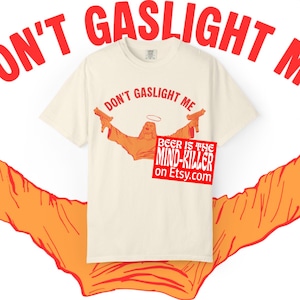 May include: Cream-colored t-shirt with the phrase "DON'T GASLIGHT ME" in red. The shirt features an orange graphic of a figure with a halo holding two guns. A red square with the text "BEER IS THE MIND-KILLER on Etsy.com" is also visible.